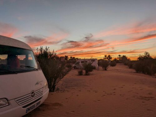 A picture of our motorhome parked in the sand dunes in the Sahara Desert at Merzouga. It is sunset with a beautiful sky and the sun reflecting off of the clouds.